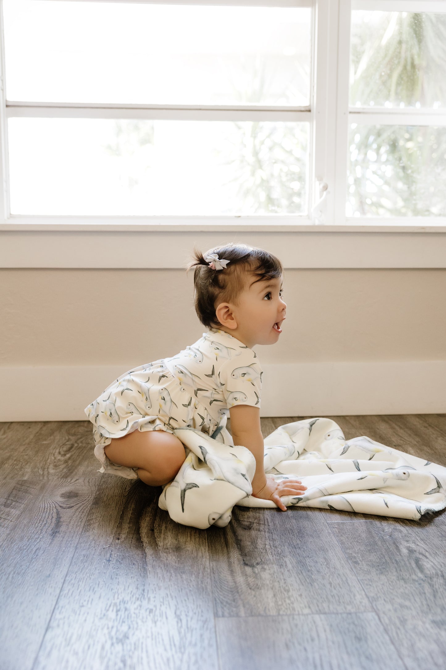 Baby sitting on a wooden floor with a yellow permit blanket, looking surprised.