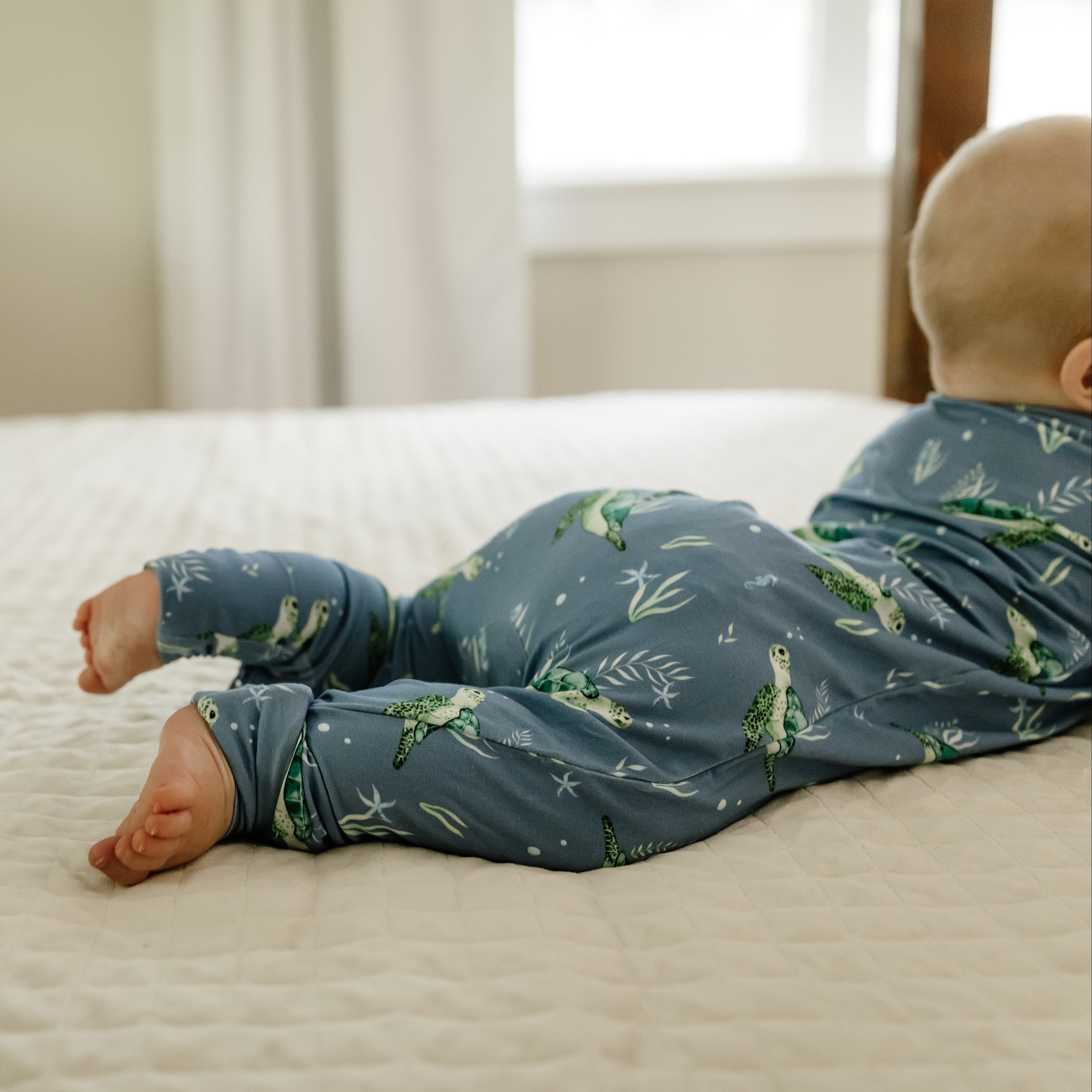 Baby in a blue onesie with green patterns lying on a bed.