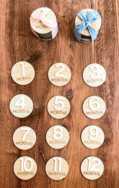 A set of wooden monthly milestone rounds for babies, engraved with numbers representing months of age. The rounds are displayed on a wooden surface, with some marked with a blue ribbon and bow.