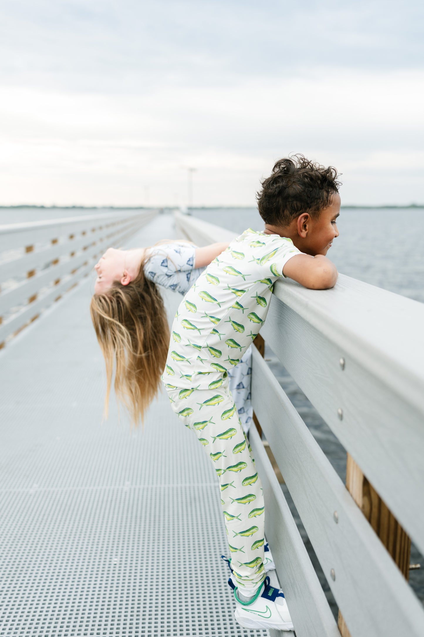 Two children wearing matching green and white patterned pajamas pose on a metal walkway overlooking water under an overcast sky.