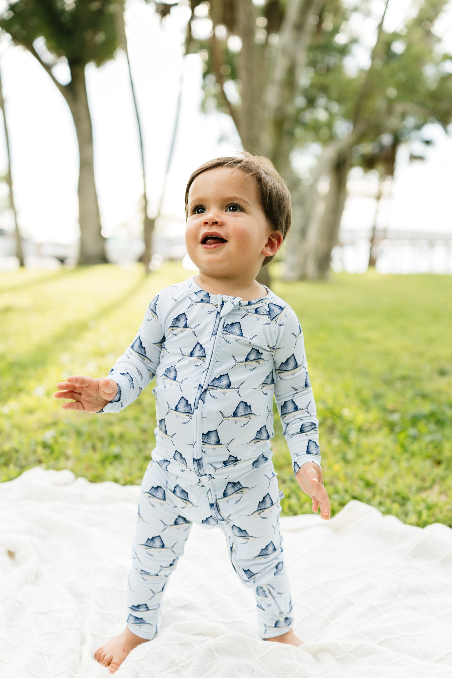 A toddler standing outdoors on a white blanket in a grassy park, wearing a light blue long-sleeve footless one-piece pajama or romper with a repeating shark print.