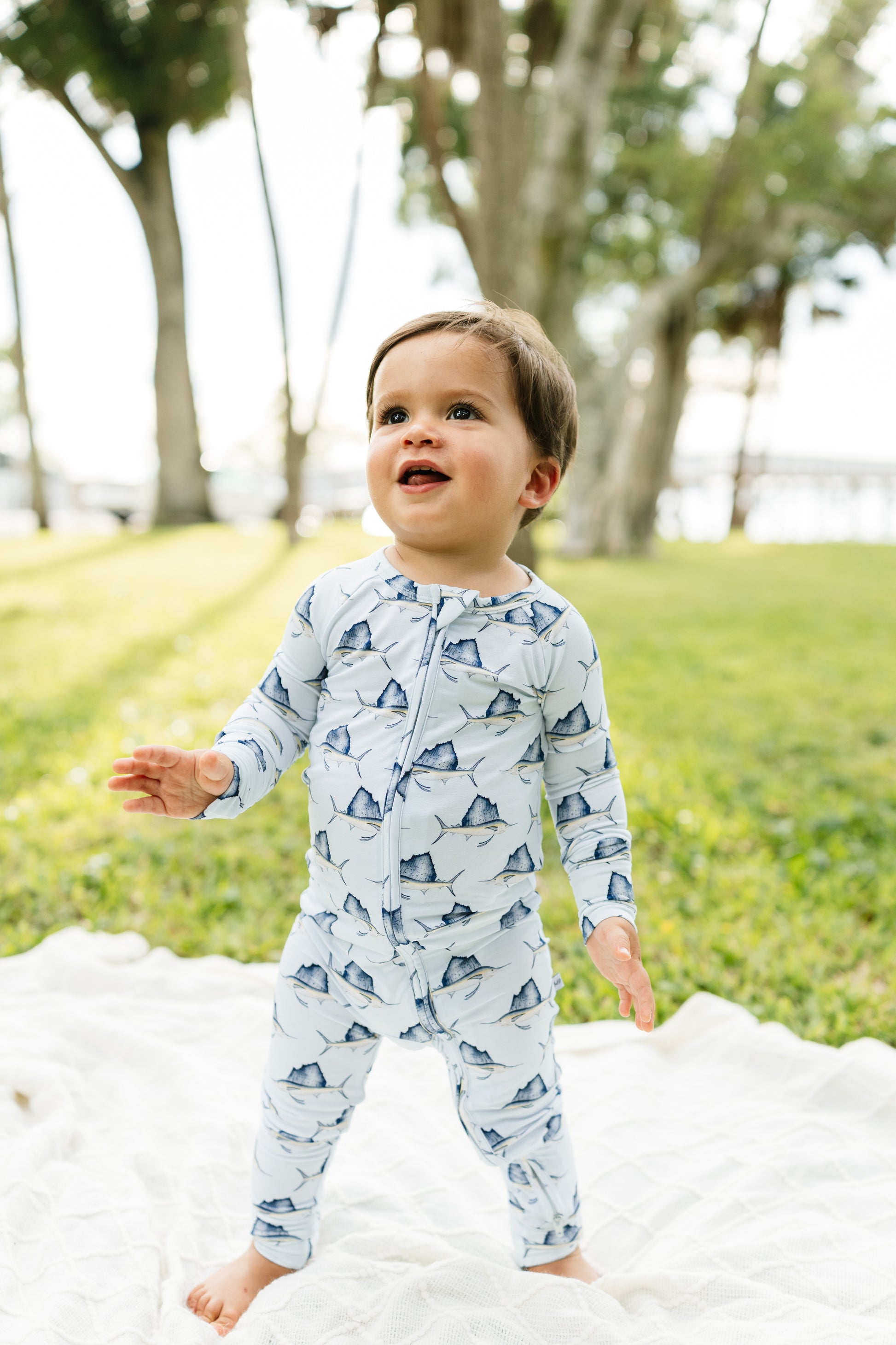 A toddler standing outdoors on a white blanket in a grassy park, wearing a light blue long-sleeve footless one-piece pajama or romper with a repeating shark print.