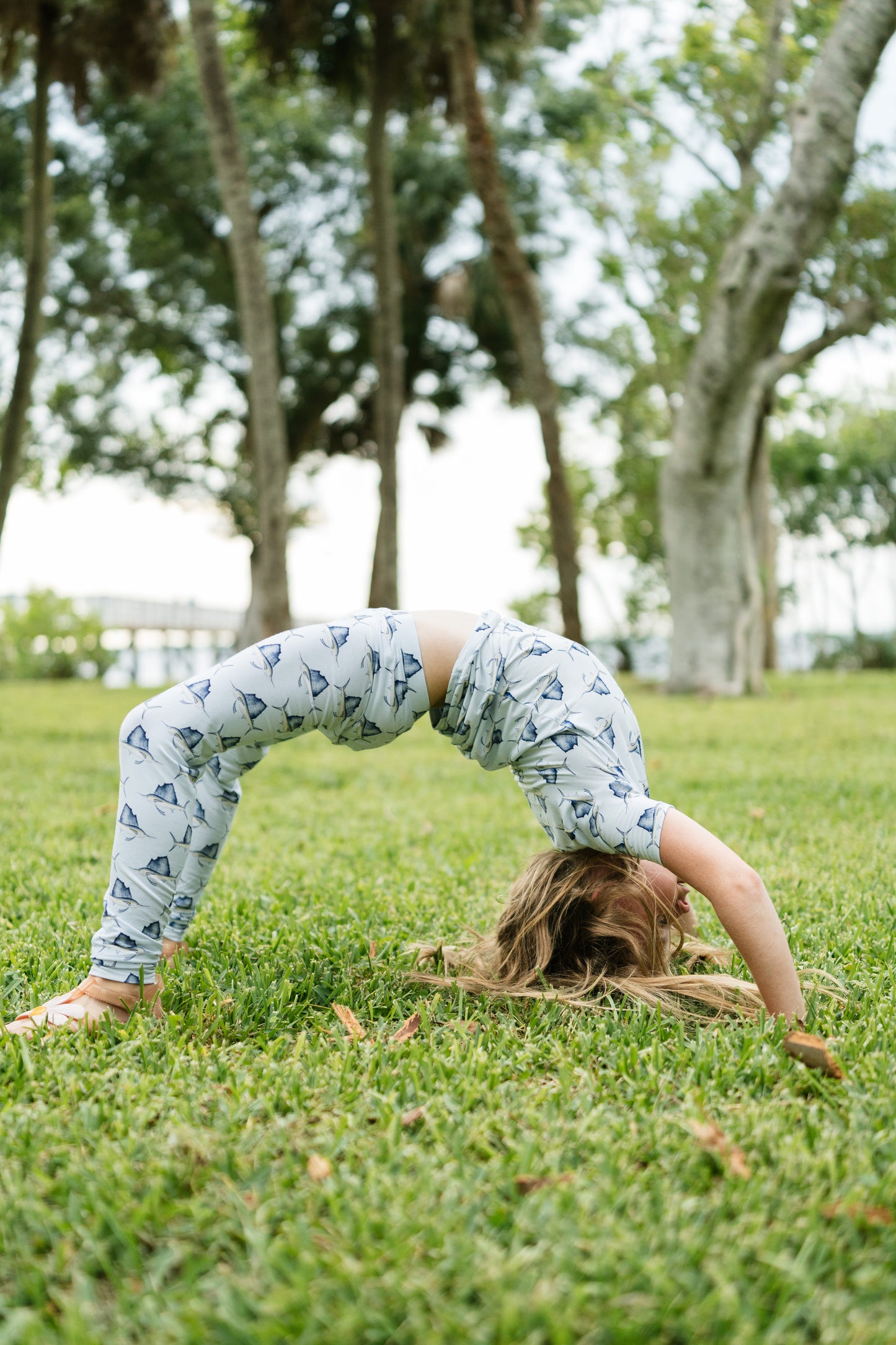 A child wearing a light blue and white patterned two-piece outfit performs a backbend on a green grassy park lawn with trees in the background.