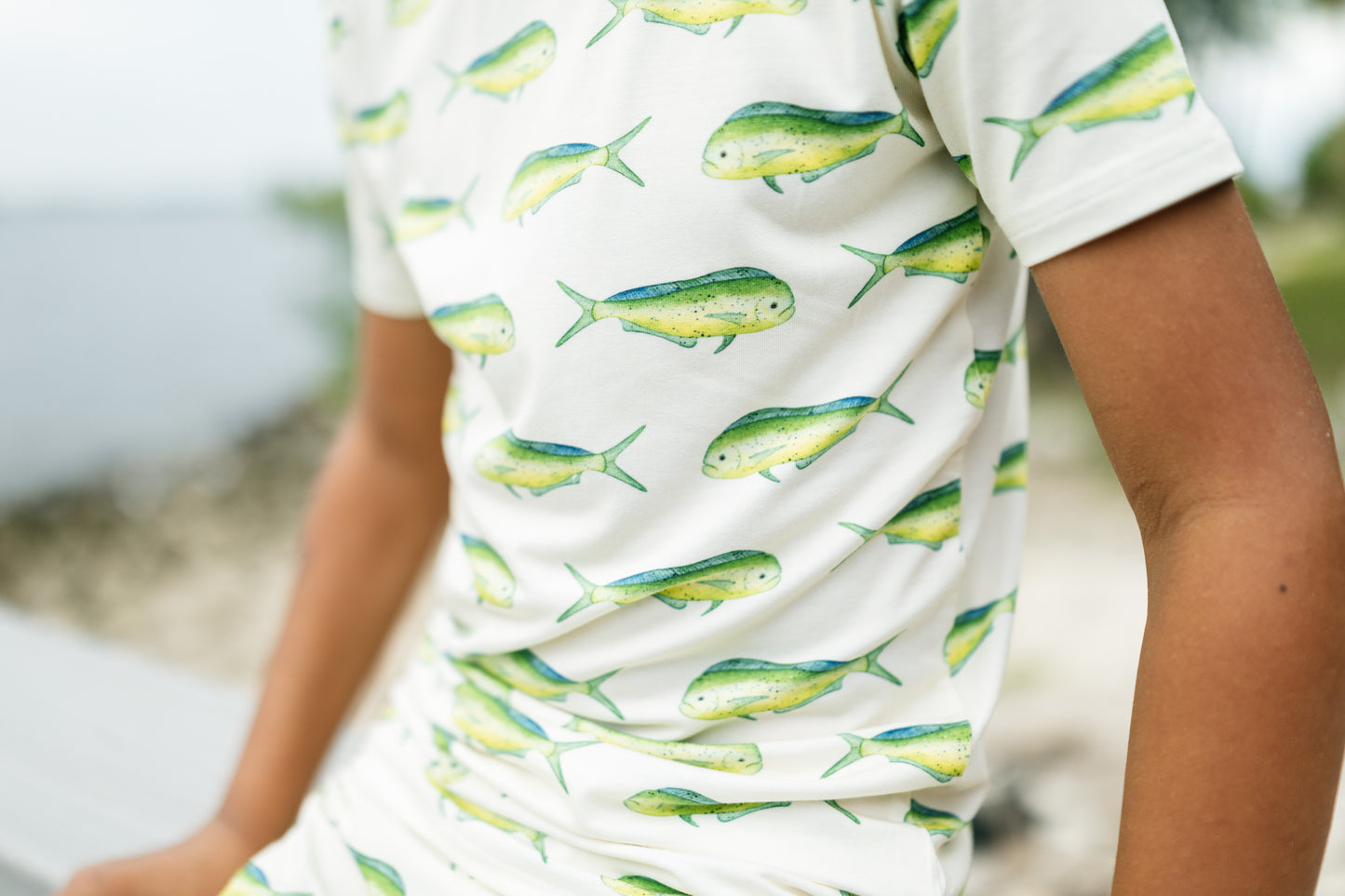 A close-up of a person wearing a white short-sleeve t-shirt with a repeating pattern of green and yellow fish, set against a blurred outdoor background.