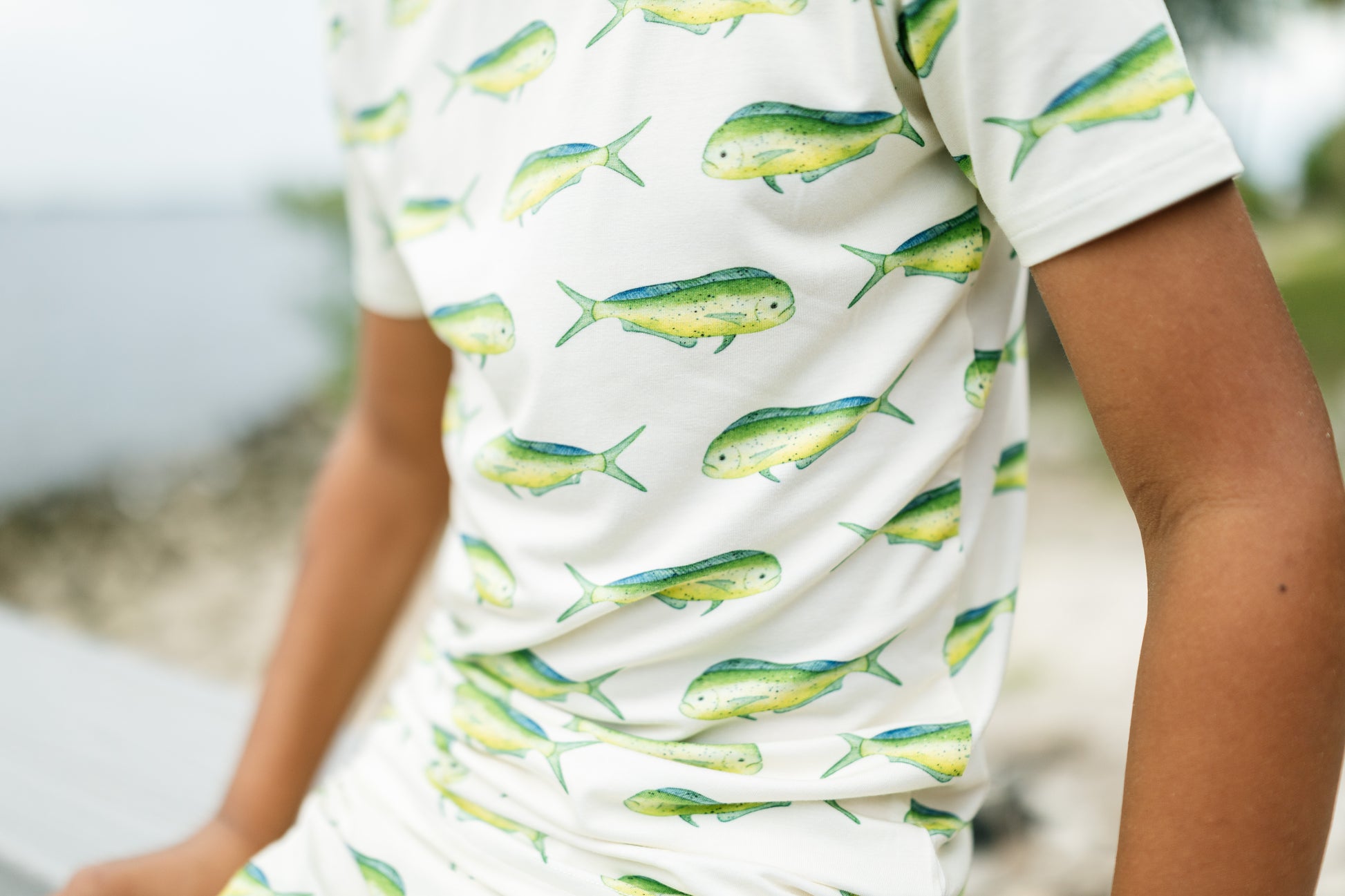 A close-up of a person wearing a white short-sleeve t-shirt with a repeating pattern of green and yellow fish, set against a blurred outdoor background.