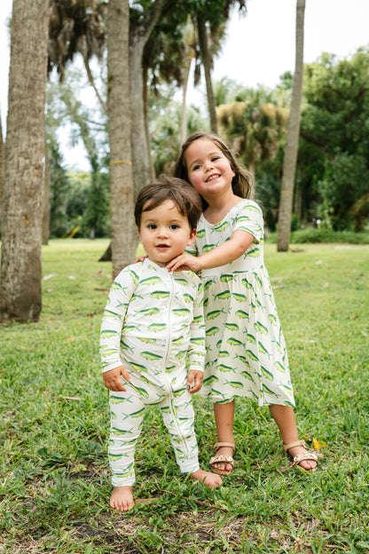 Two children standing outdoors on grass in front of palm trees, wearing matching light green and white long-sleeve outfits with a repeating leaf print.
