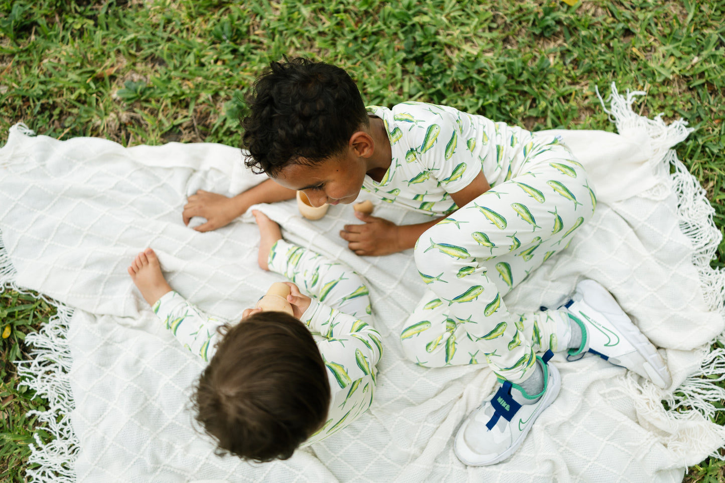 Two children wearing matching white long-sleeve pajamas with a green leaf print, sitting on a white blanket on grass outdoors.