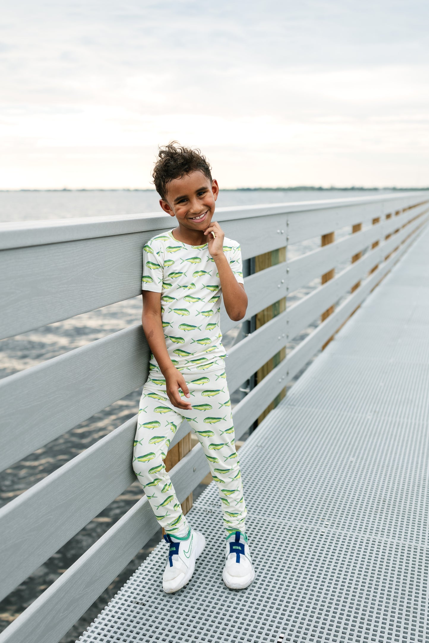 A young boy wearing a matching two-piece pajama set with a green alligator print on a white background, leaning against a gray wooden railing on a metal walkway.