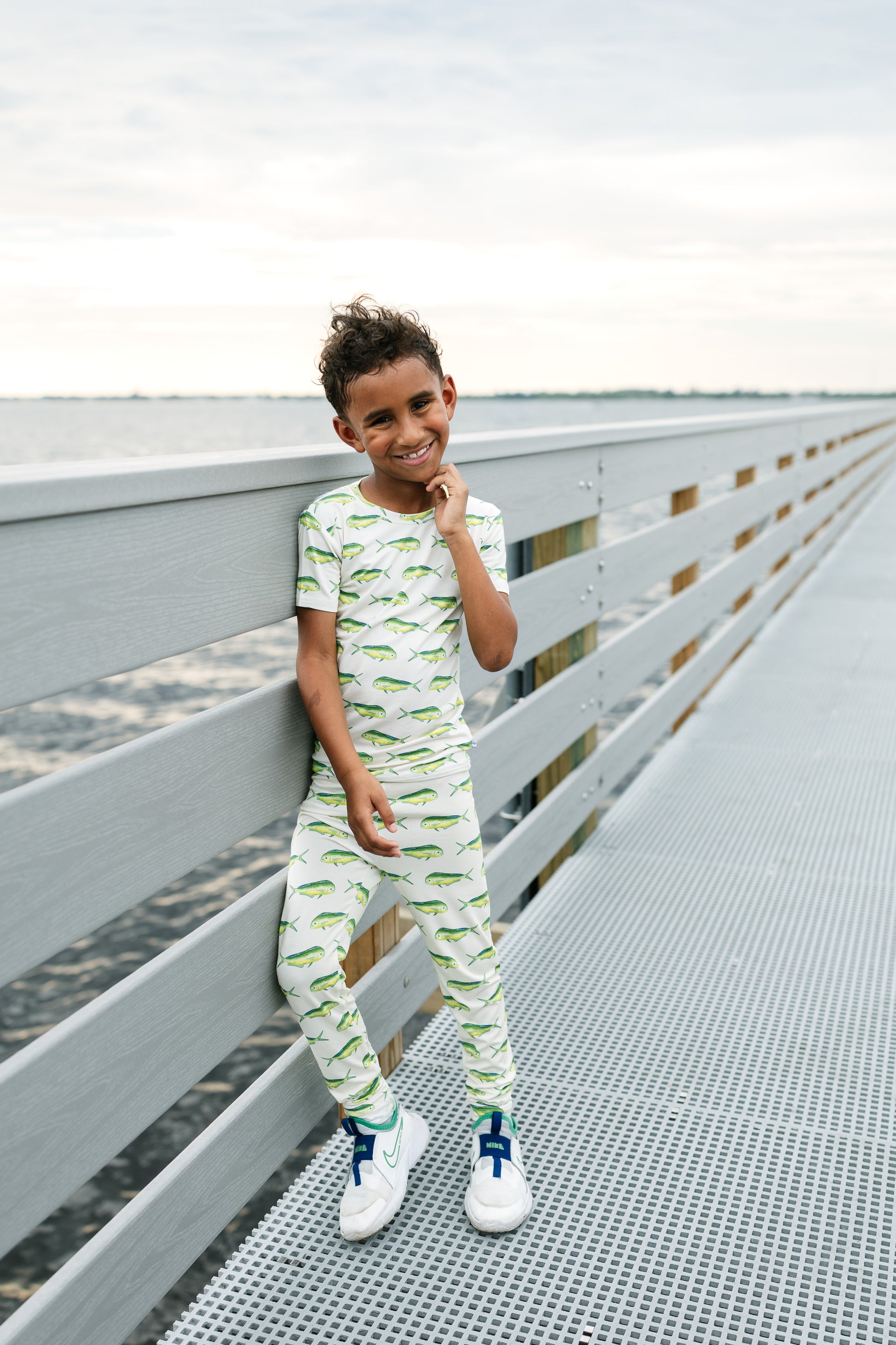 A young boy wearing a matching two-piece pajama set with a green alligator print on a white background, leaning against a gray wooden railing on a metal walkway.