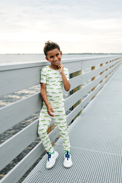 A young boy wearing a matching two-piece pajama set with a green alligator print on a white background, leaning against a gray wooden railing on a metal walkway.