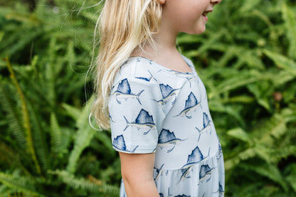 A young girl with blonde hair is shown in profile wearing a light blue dress with a navy blue sailfish print, set against a background of green ferns.