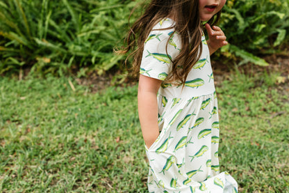 A young girl wearing a white short-sleeve dress with a green and yellow fish print, standing outdoors on a grassy lawn with green foliage in the background.