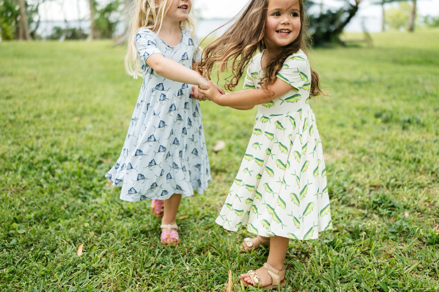 Two young girls holding hands and smiling while running on a green grassy lawn. The girl on the left wears a light blue dress with a small blue pattern, and the girl on the right wears a white dress with green leaf prints.