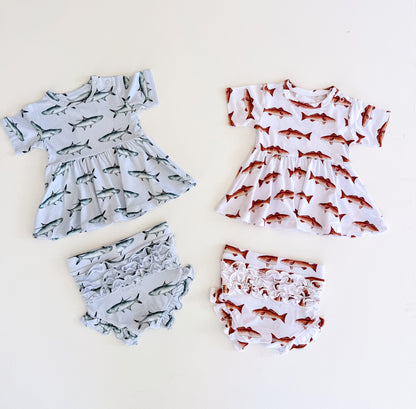 Flat lay of two peplum and ruffle bloomer sets one with light blue background with Tarpon one with white background with red fish both have a snap on the left shoulder and short sleeves