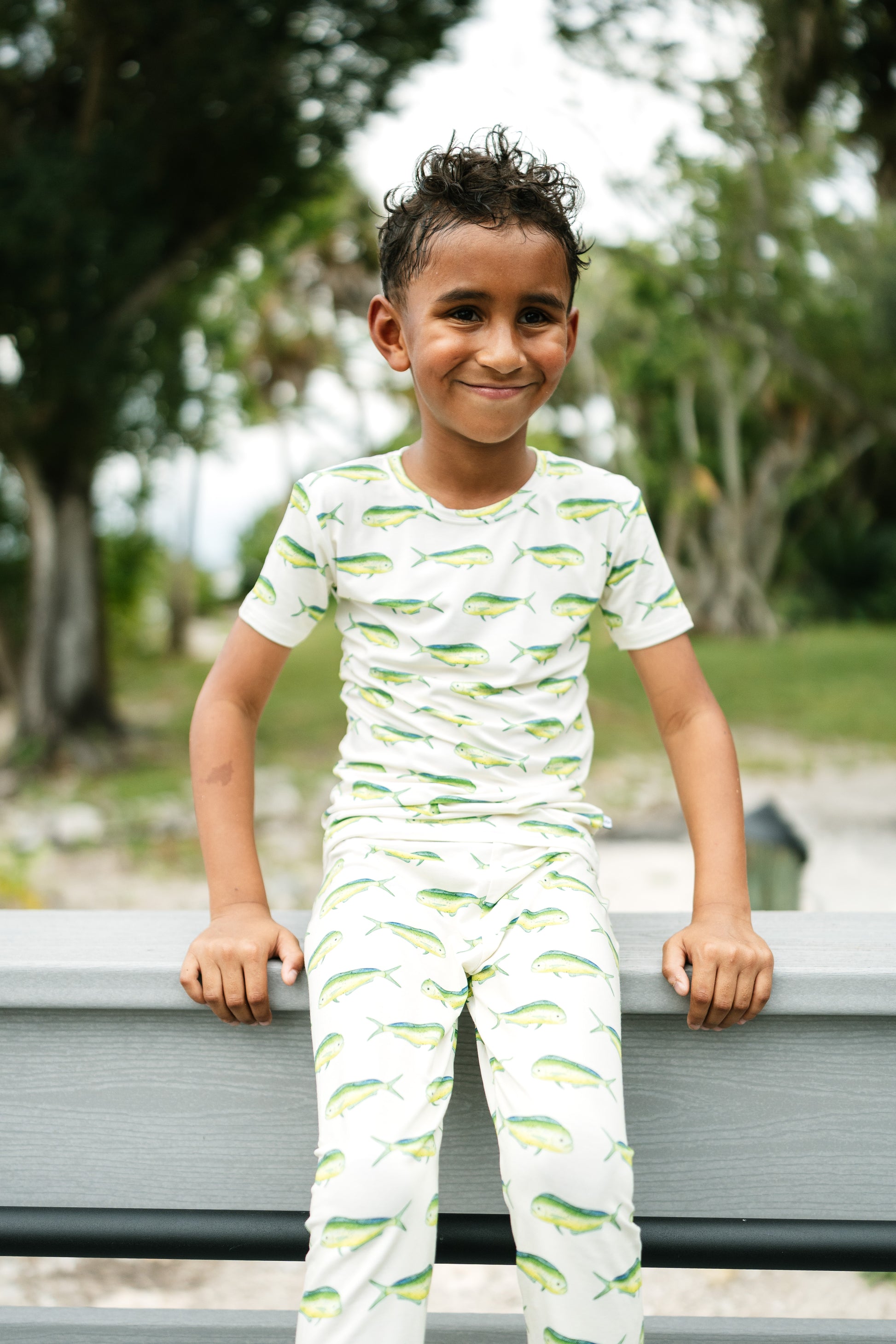 A young boy smiling while wearing a matching two-piece pajama set with a green fish pattern on a white background, leaning on a gray bench in an outdoor park setting.