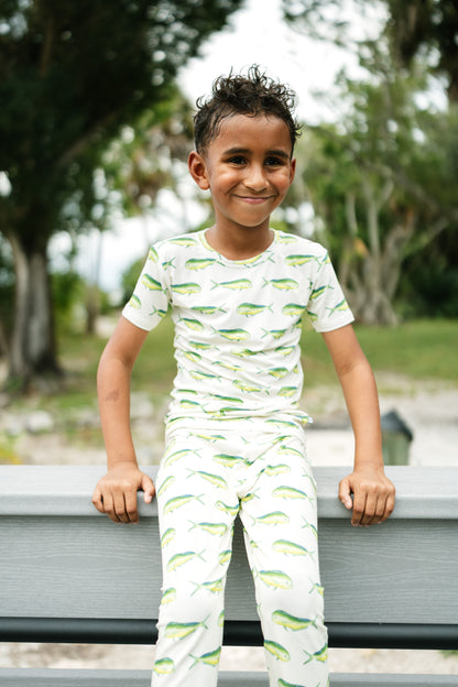 A young boy smiling while wearing a matching two-piece pajama set with a green fish pattern on a white background, leaning on a gray bench in an outdoor park setting.