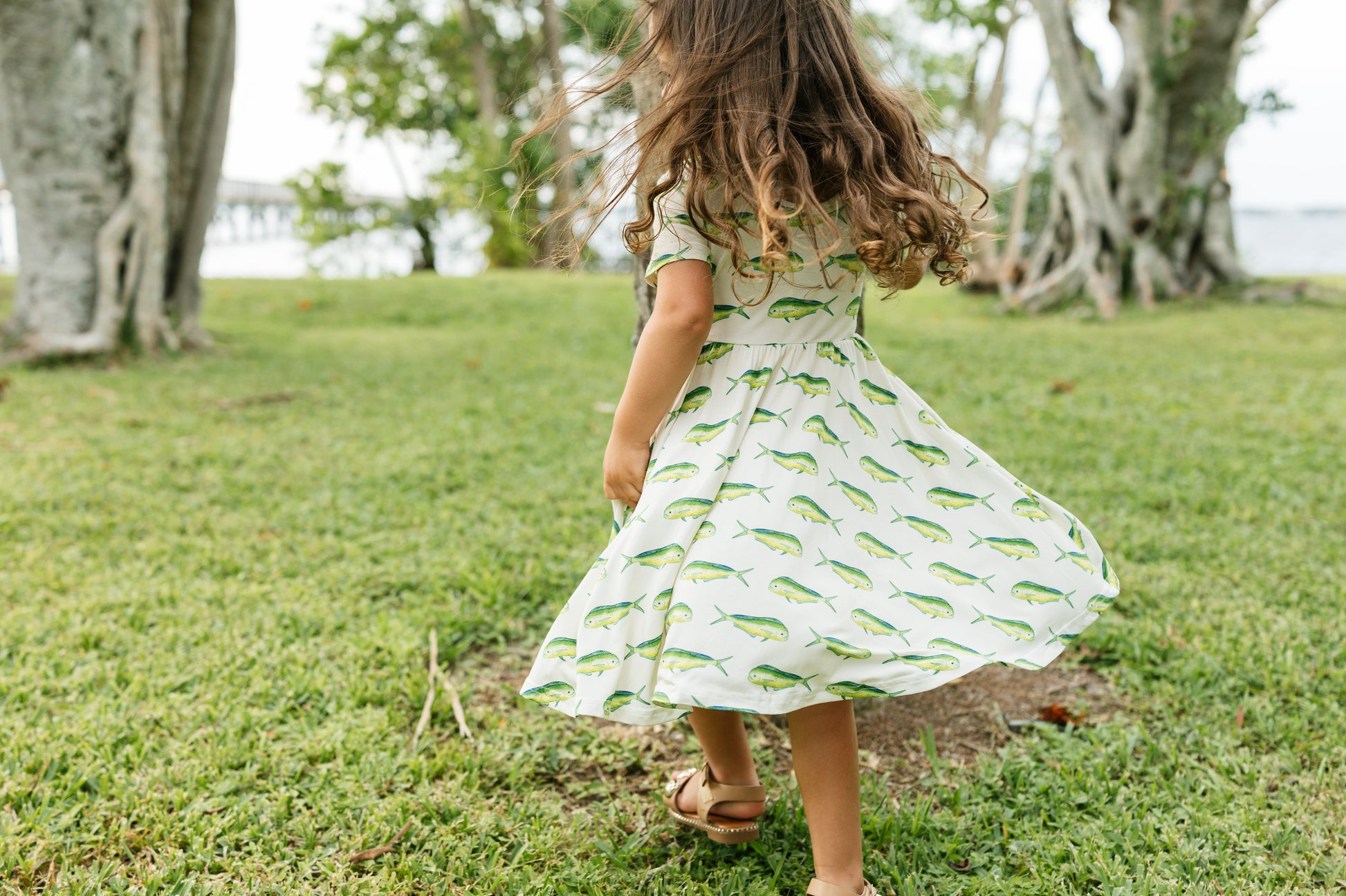 A young girl with long brown hair is seen from behind, wearing a white short-sleeve dress with a green fish pattern, twirling on a grassy lawn outdoors.