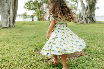A young girl with long brown hair is seen from behind, wearing a white short-sleeve dress with a green fish pattern, twirling on a grassy lawn outdoors.