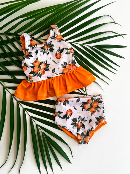 Children's swimsuit set with orange and floral pattern on a white background with palm leaves.