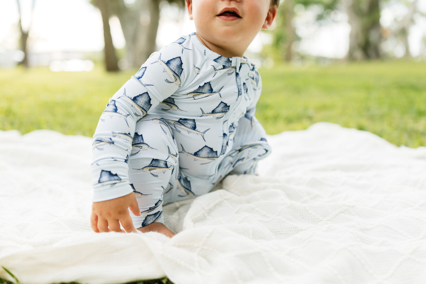 A baby sitting on a white blanket in a park, wearing a long-sleeve light blue and white shark print pajama or romper.