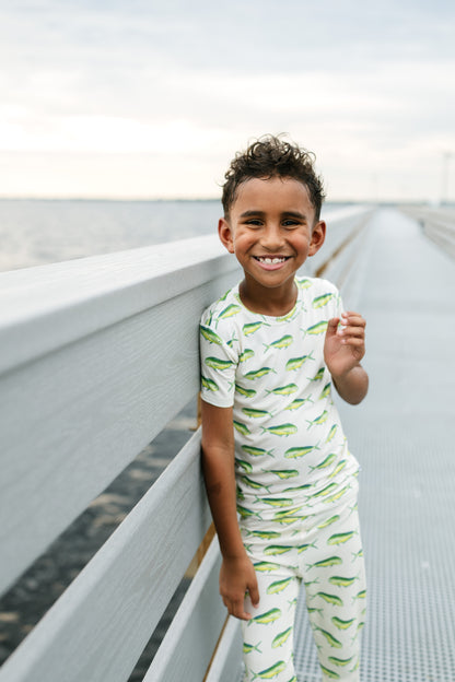 A smiling young boy stands on a boardwalk railing wearing a white short-sleeve pajama set with a green fish print. The background shows a soft-focus outdoor scene with water and sky.