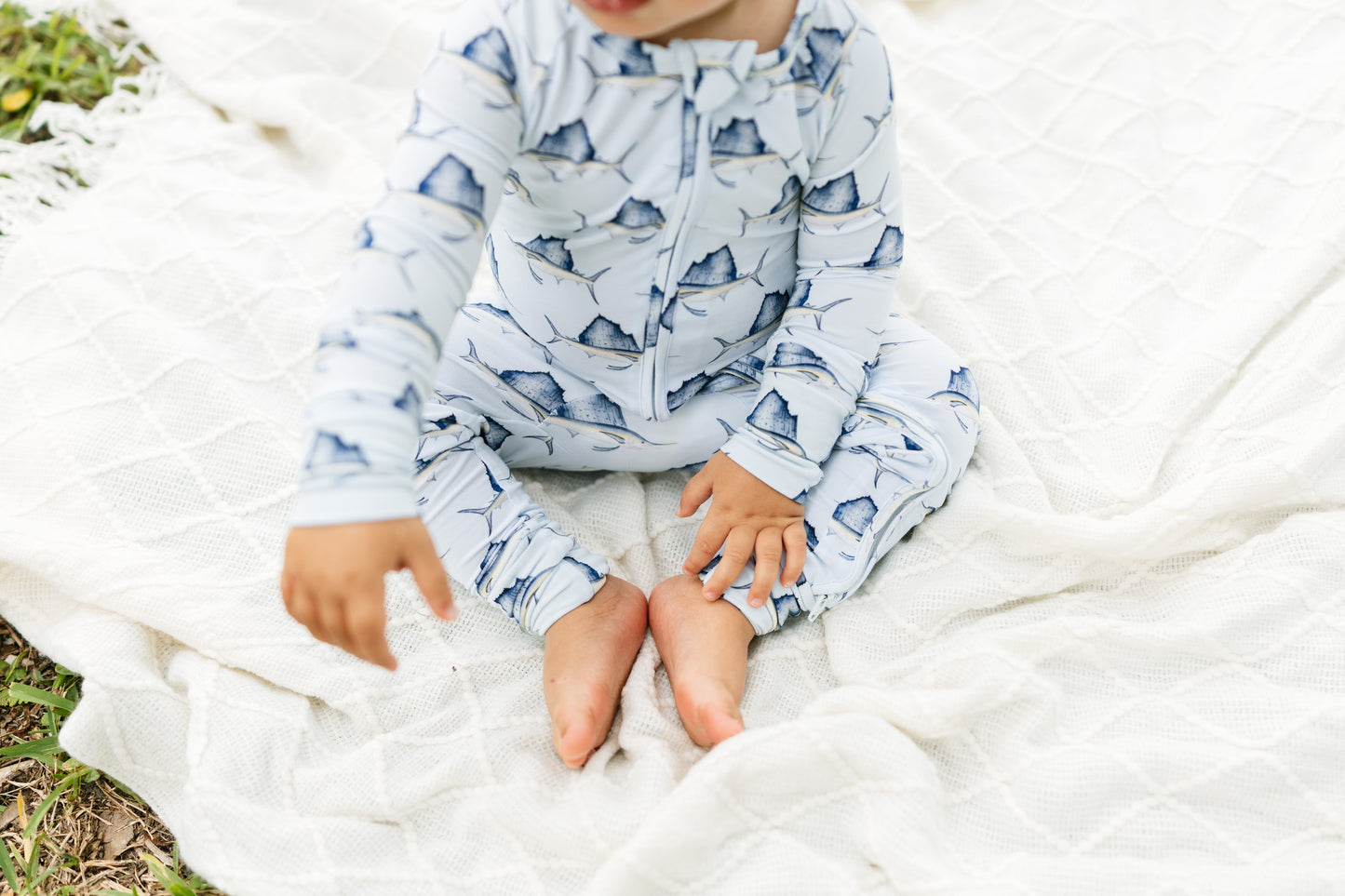 A baby sitting on a white quilted blanket outdoors, wearing a light blue long-sleeve zip-up sleeper with a dark blue mountain pattern.