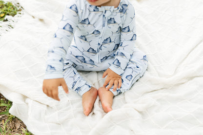 A baby sitting on a white quilted blanket outdoors, wearing a light blue long-sleeve zip-up sleeper with a dark blue mountain pattern.