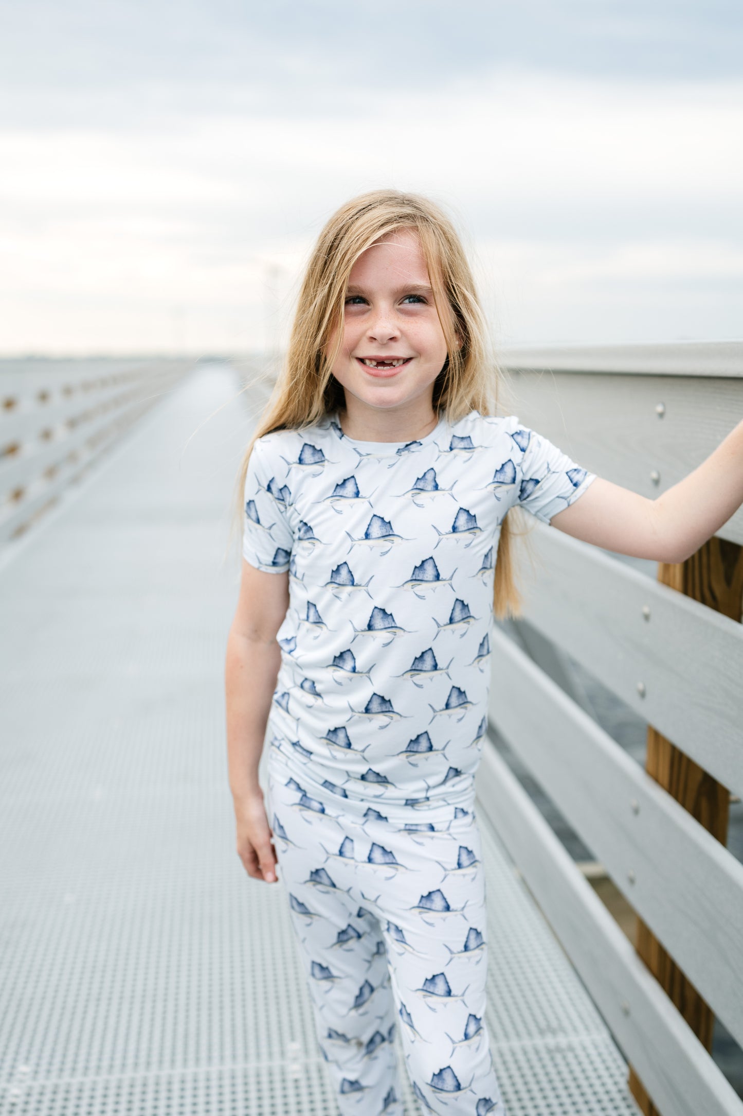 A young girl with blonde hair stands on a metal walkway wearing a matching short-sleeve pajama set with a blue sailboat pattern on a white background.