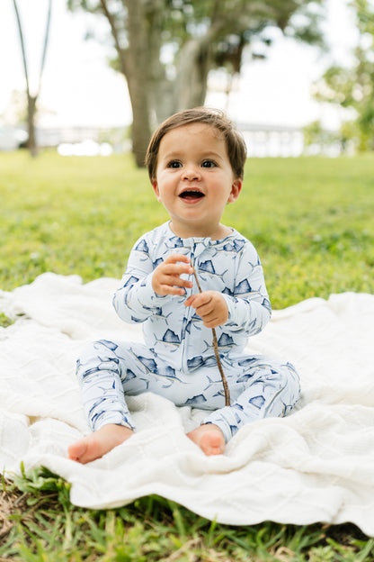 A baby sitting outdoors on a white blanket in a grassy park, wearing a light blue and white patterned long-sleeve romper and holding a small twig.
