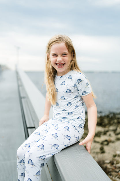 A young girl with blonde hair sits smiling on a gray wooden boardwalk railing, wearing a matching white short-sleeve pajama top and pants set with a blue fish print. The background shows a soft-focus outdoor coastal scene with water and sky.