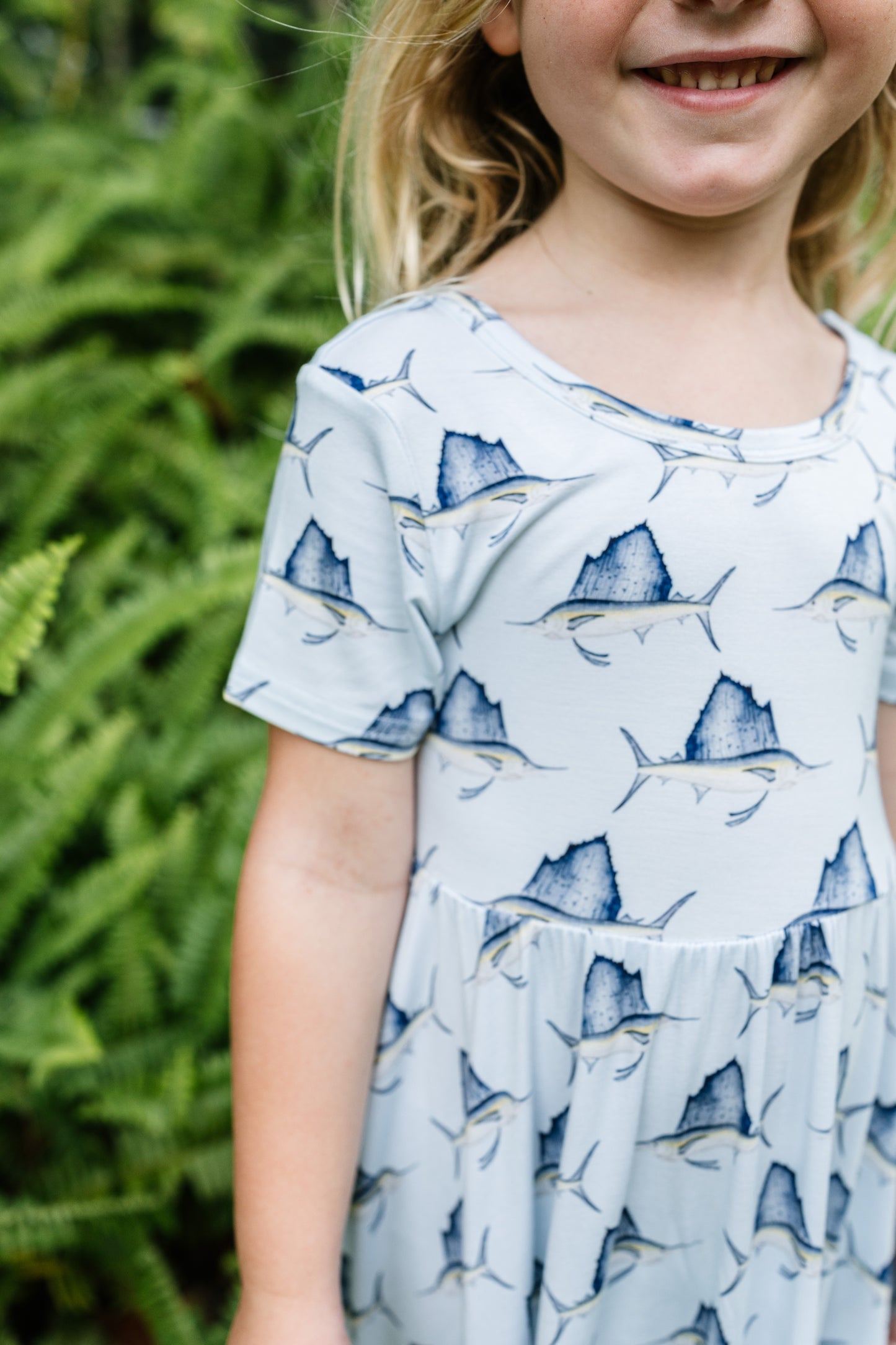 A young girl wearing a light blue short-sleeve dress with a blue and gray marlin print, standing outdoors in front of a green fern background.
