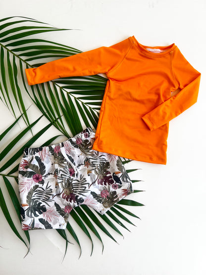 Orange long-sleeve rashguard swim shirt and floral shorts on a white background with green leaves.
