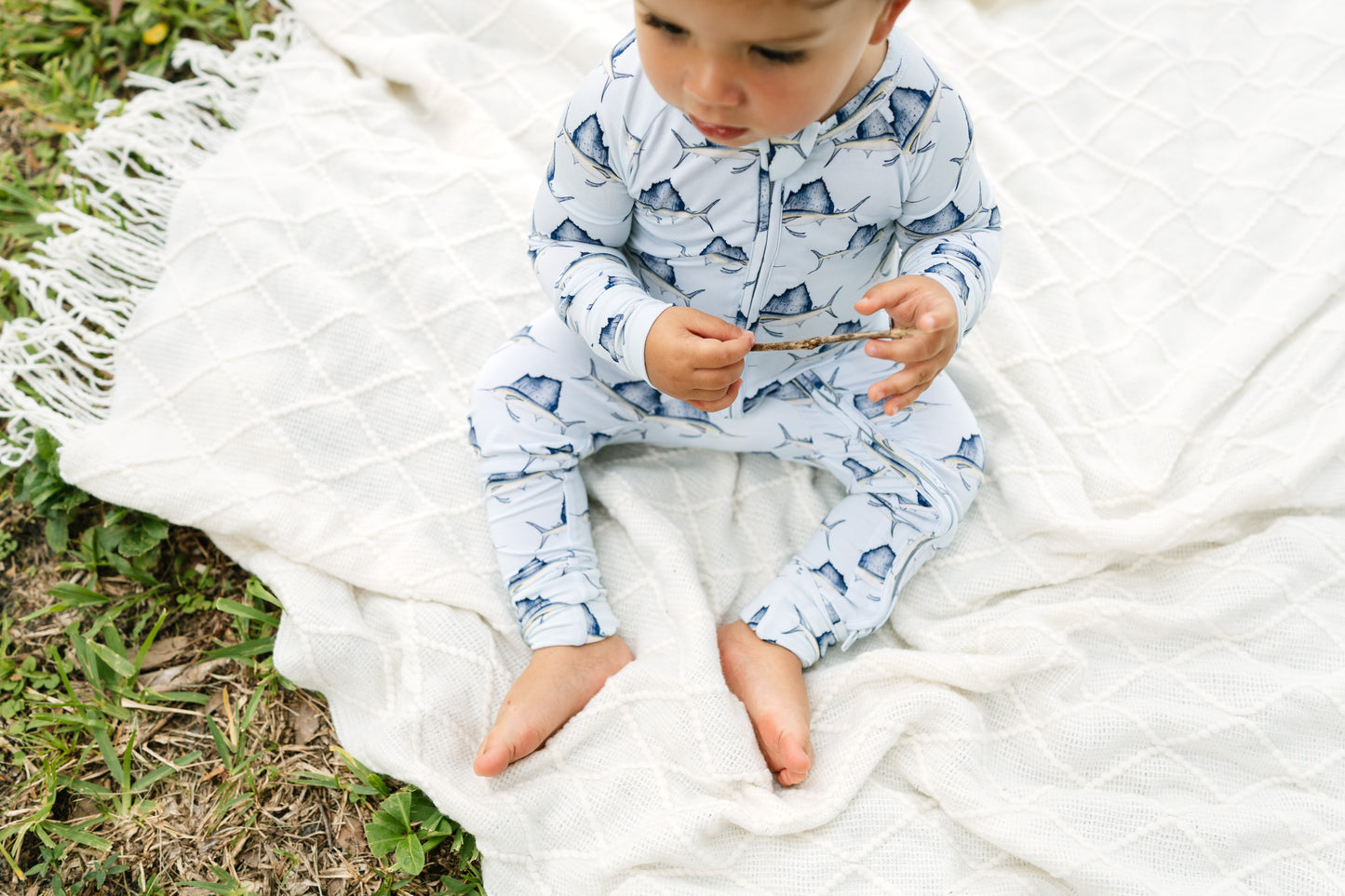A baby sitting barefoot on a white quilted blanket outdoors, wearing a light blue and white patterned zip-up one-piece pajama.