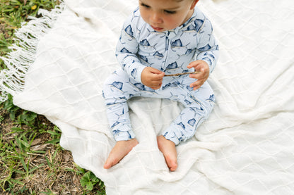 A baby sitting barefoot on a white quilted blanket outdoors, wearing a light blue and white patterned zip-up one-piece pajama.