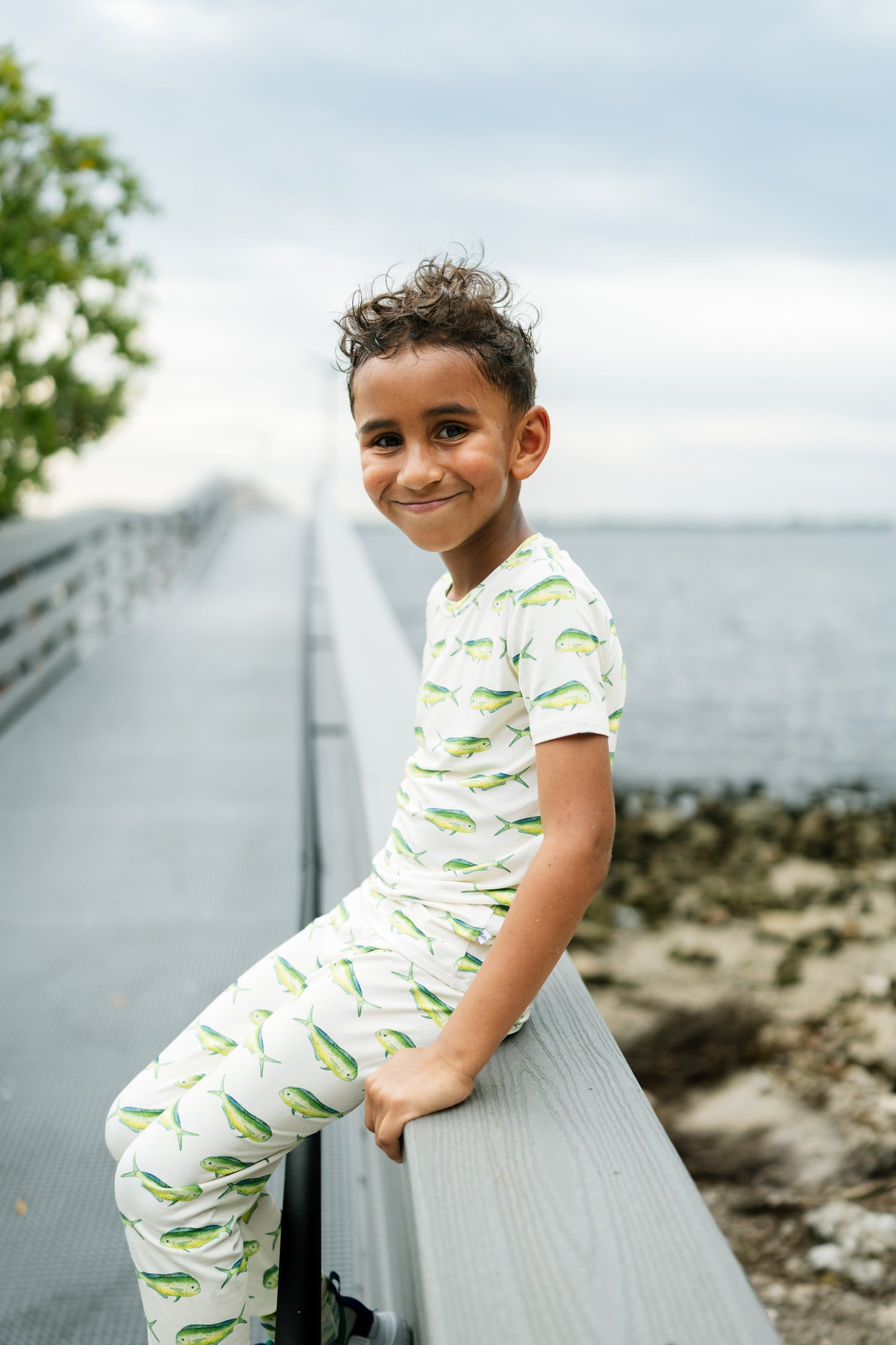 A young boy wearing a white short-sleeve pajama set with a green fish pattern, leaning against a gray wooden boardwalk railing outdoors.