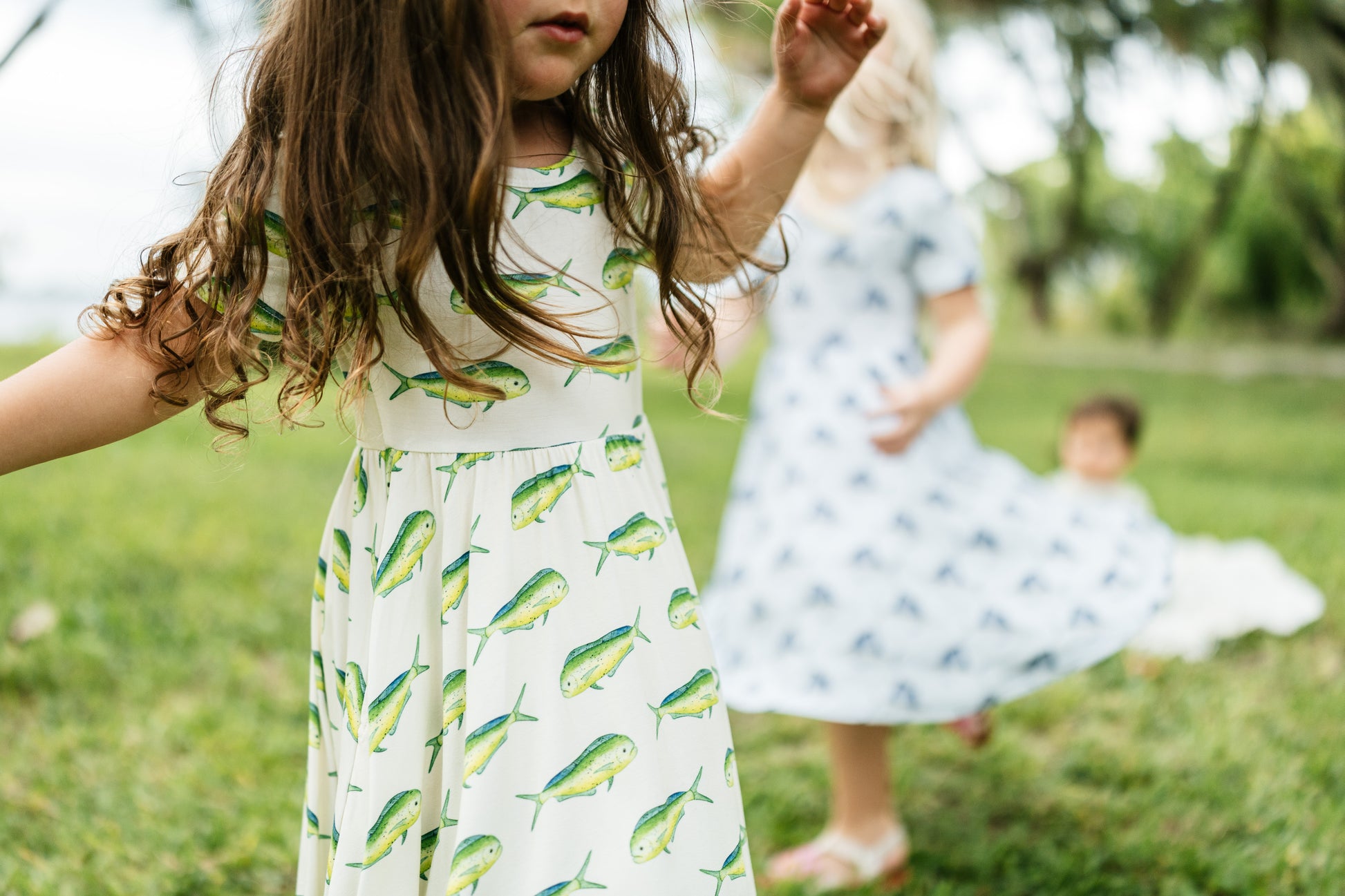 A young girl in a white dress with a green and yellow mahi-mahi fish print stands in a grassy park, with another child in a light blue patterned dress blurred in the background.