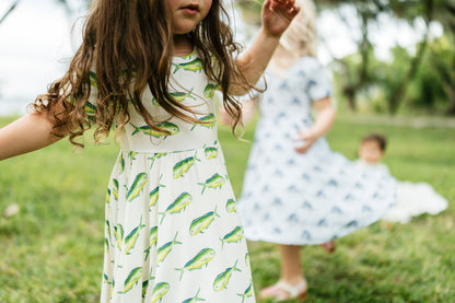 A young girl in a white dress with a green and yellow mahi-mahi fish print stands in a grassy park, with another child in a light blue patterned dress blurred in the background.
