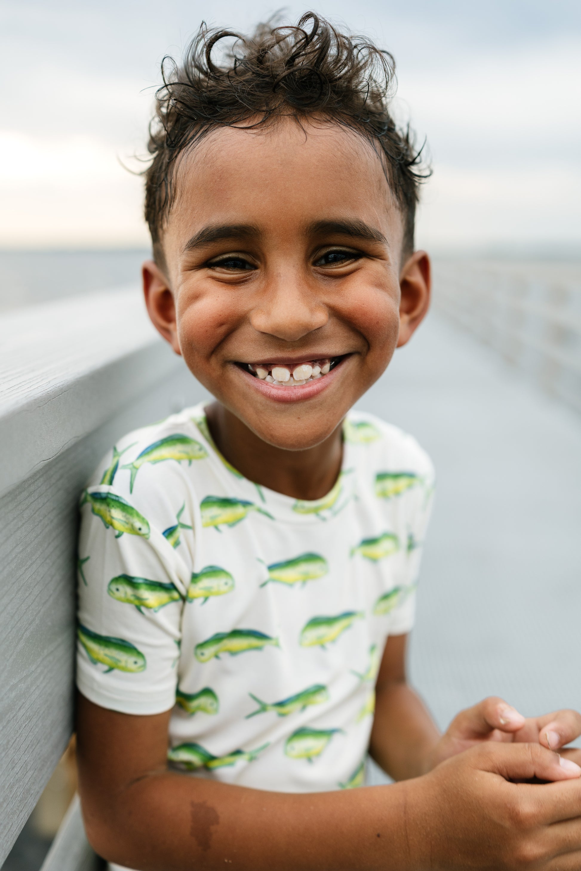 A smiling young boy with curly dark hair leans against a gray railing on a pier, wearing a white short-sleeve t-shirt with a green and yellow fish pattern.
