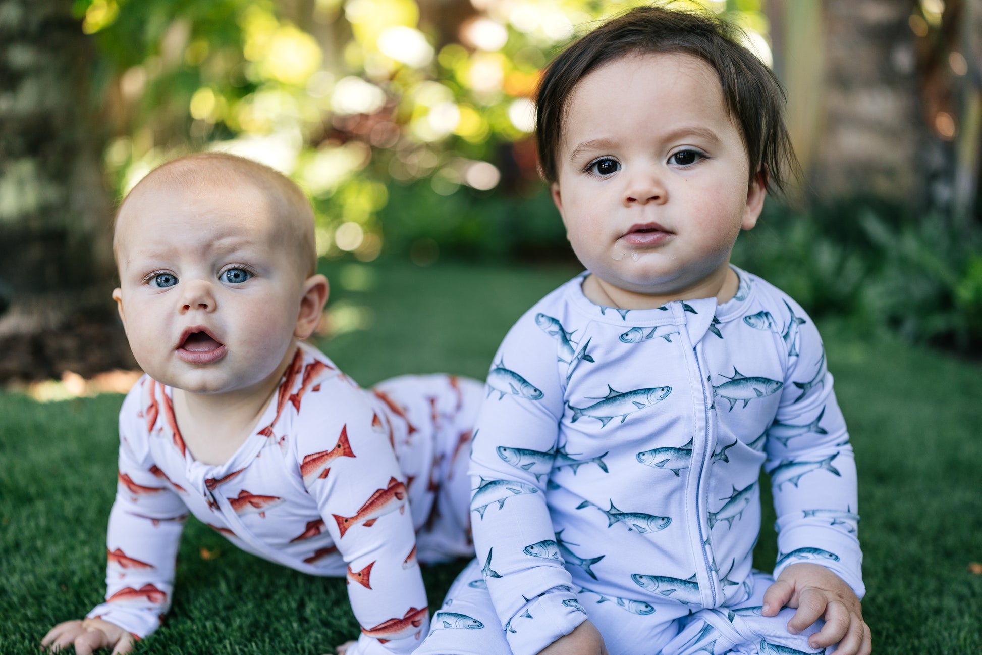 Two babies wearing tarpon and redfish outfits sitting on grass with a blurred natural background