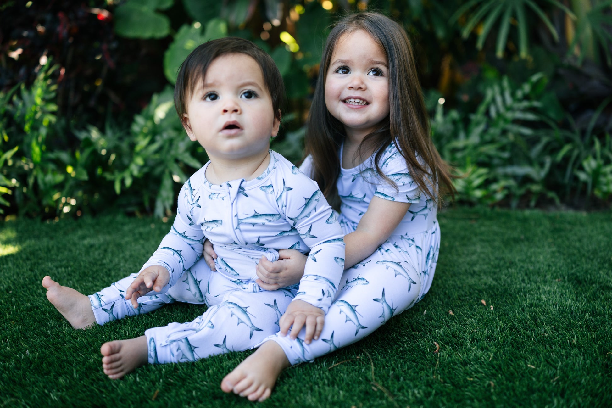 Two children in matching pajamas sitting on grass with a natural background