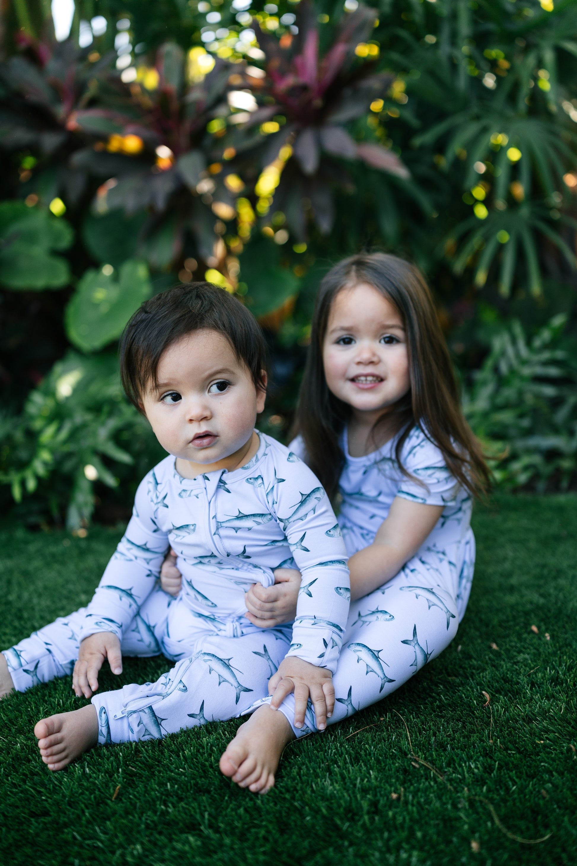 Two children in matching pajamas lying on grass with plants in the background