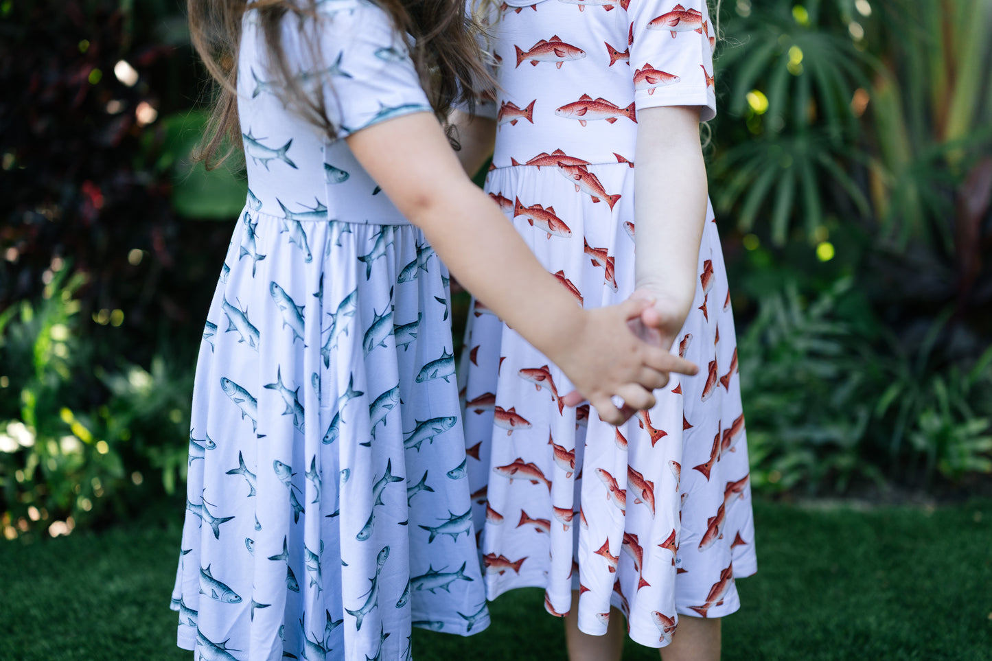 Two children holding hands wearing dresses with fish patterns in a garden setting.