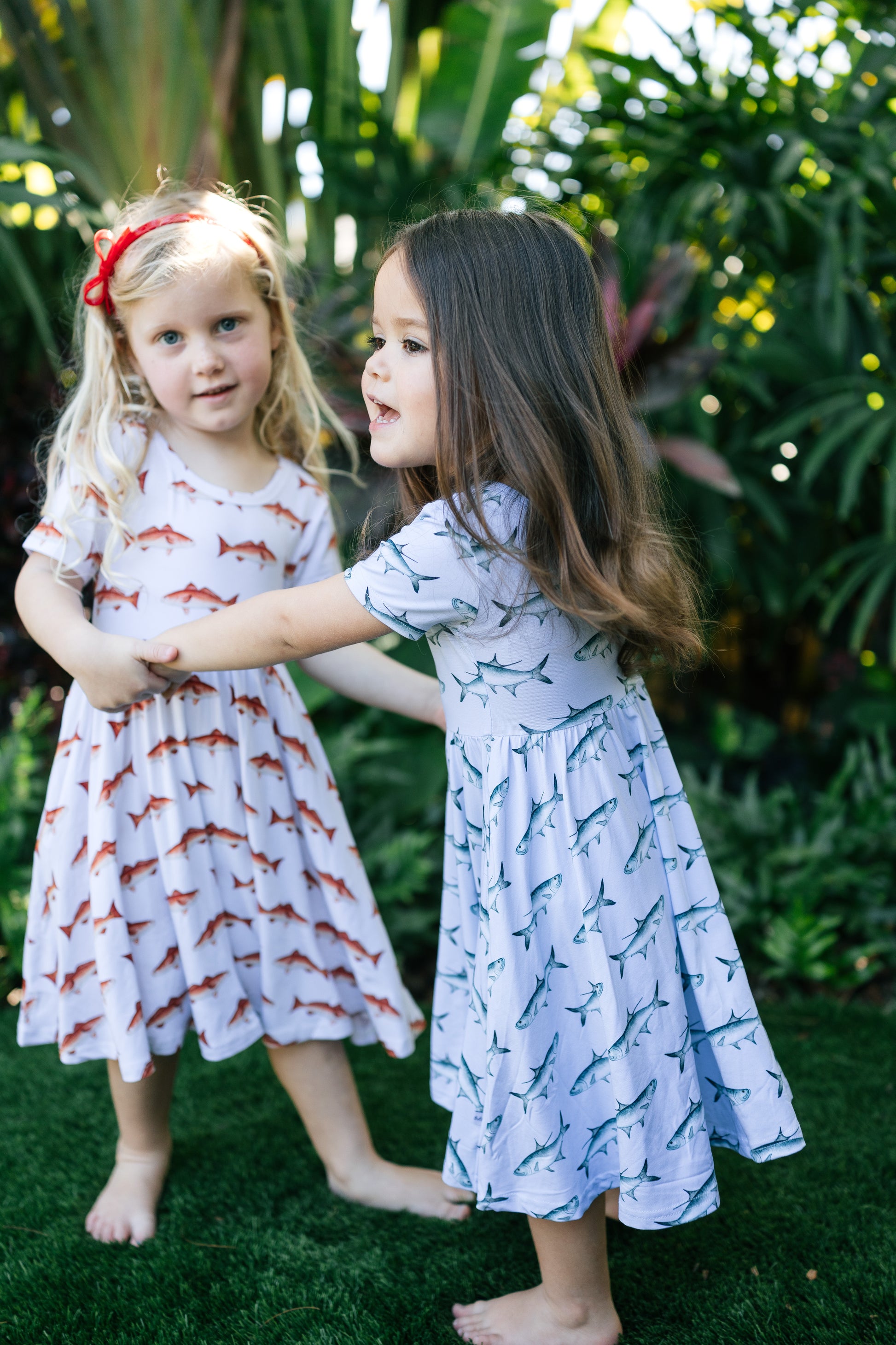 Two children in patterned dresses standing outdoors with greenery in the background
