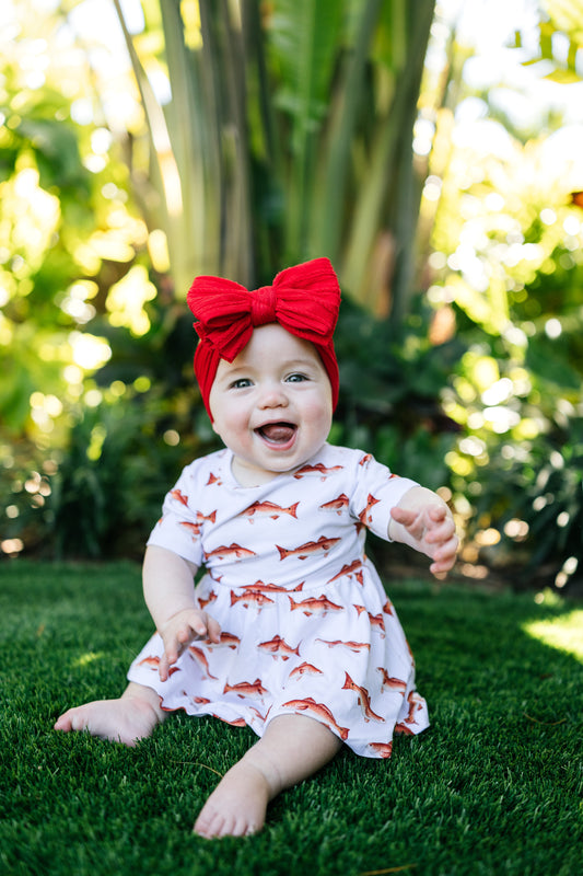 baby in redfish pattern sitting up smiling outdoors with a big red bow