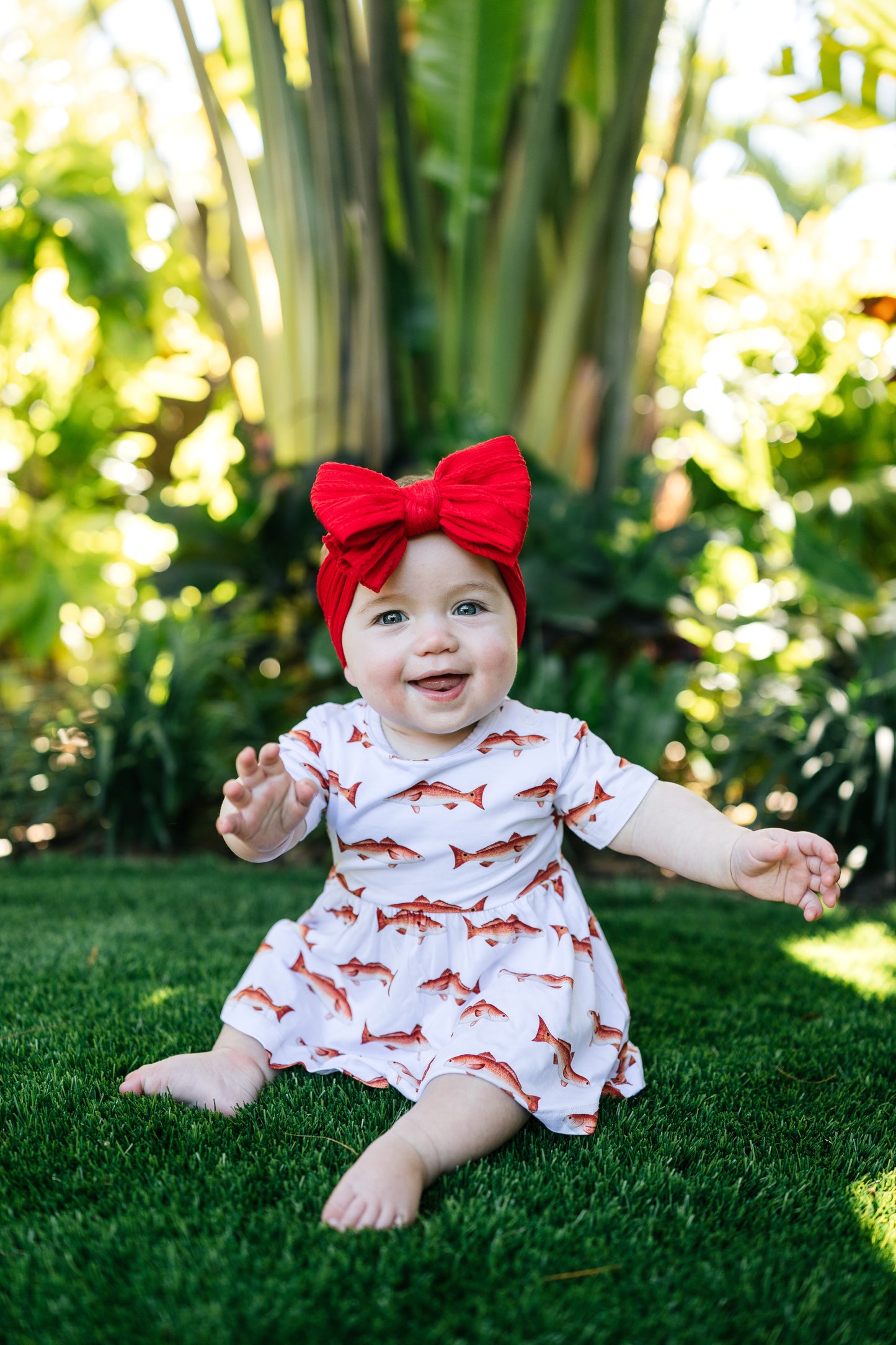 baby in redfish pattern sitting up smiling outdoors with a big red bow