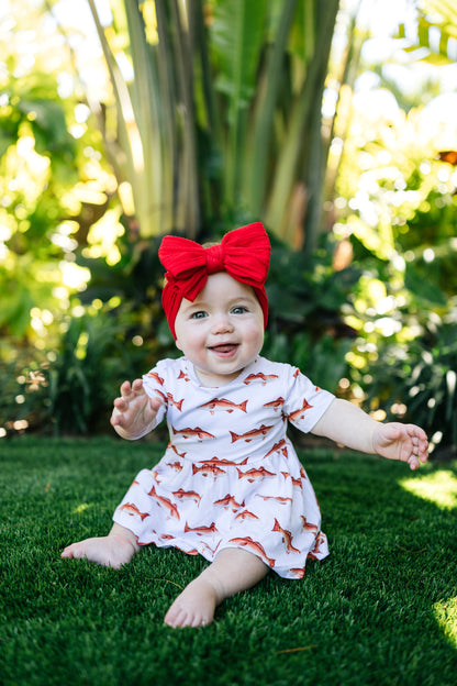 baby in redfish pattern sitting up smiling outdoors with a big red bow