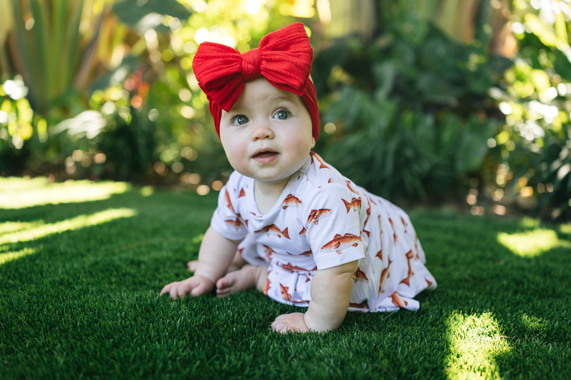baby in redfish pattern sitting up smiling outdoors with a big red bow
