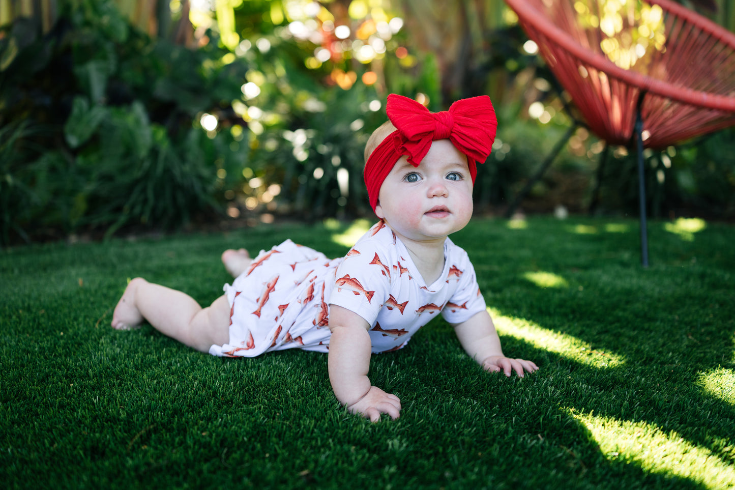 Baby wearing a red bow headband and white outfit with redfish , lying on grass.