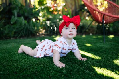 Baby wearing a red bow headband and white outfit with redfish , lying on grass.