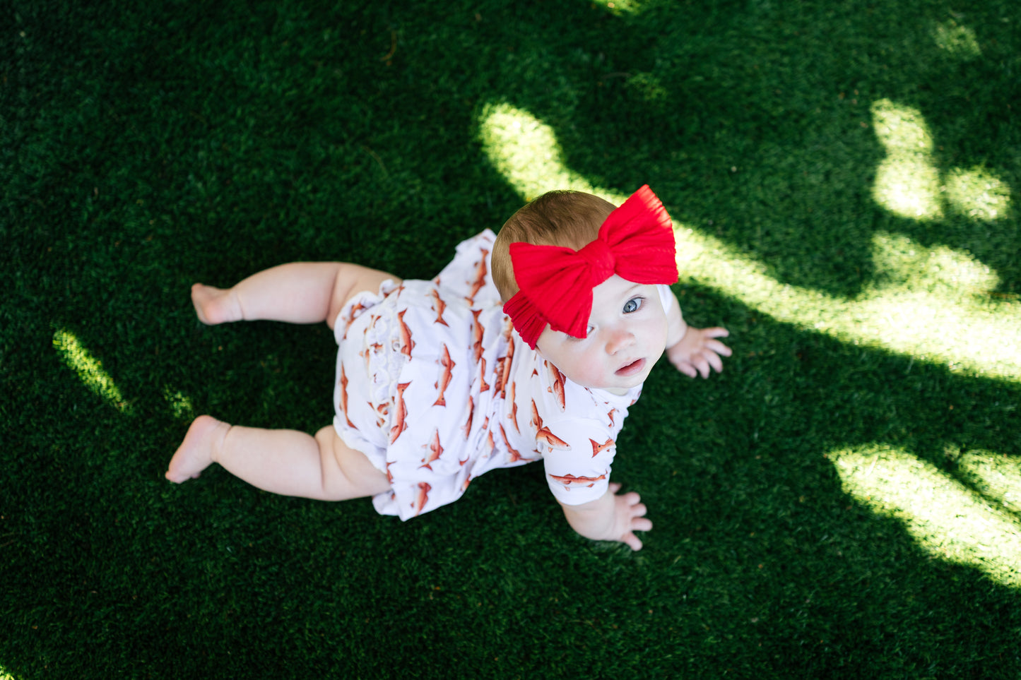 Baby crawling on grass wearing a red bow and white outfit with redfish patterns